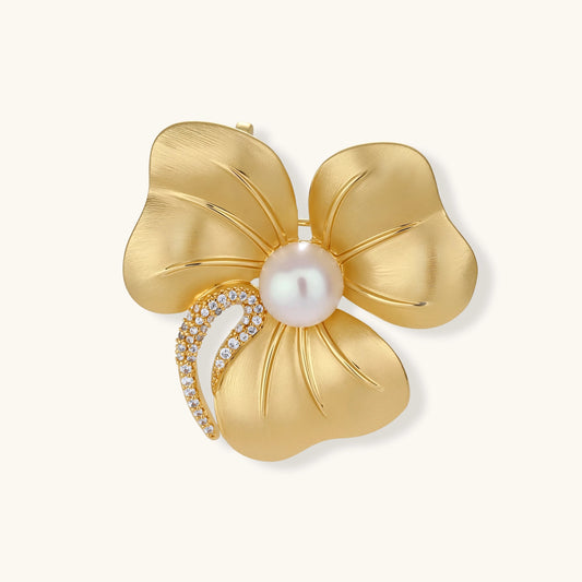 Gold-tone three-petal flower brooch with a brushed finish, round white bead center and zirconia crescent on a cream background.