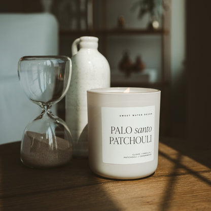Palo Santo Patchouli candle in a tan jar on a table beside a glass and ceramic vase.