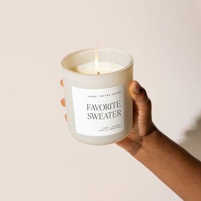 Lit Favorite Sweater candle in a tan jar held against a pale background.