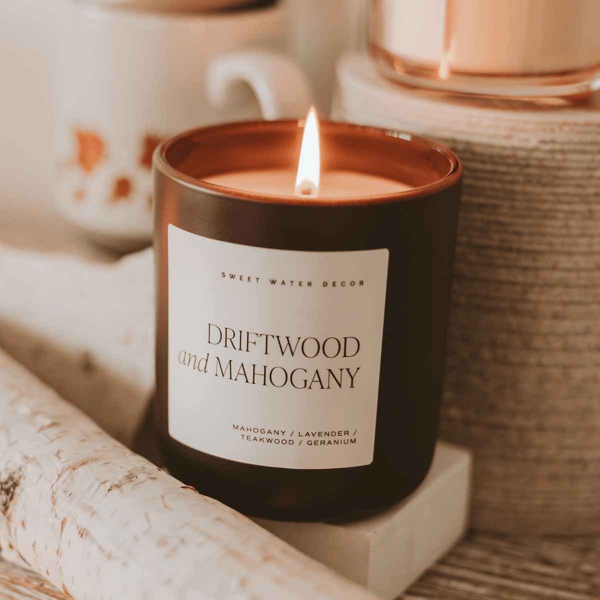 Lit Driftwood and Mahogany candle in a brown jar beside a mug and stacked books.
