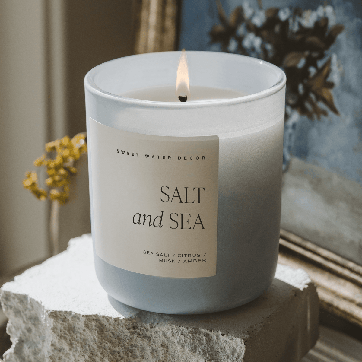 Lit Salt and Sea candle in a blue jar on stacked stone beside a window.