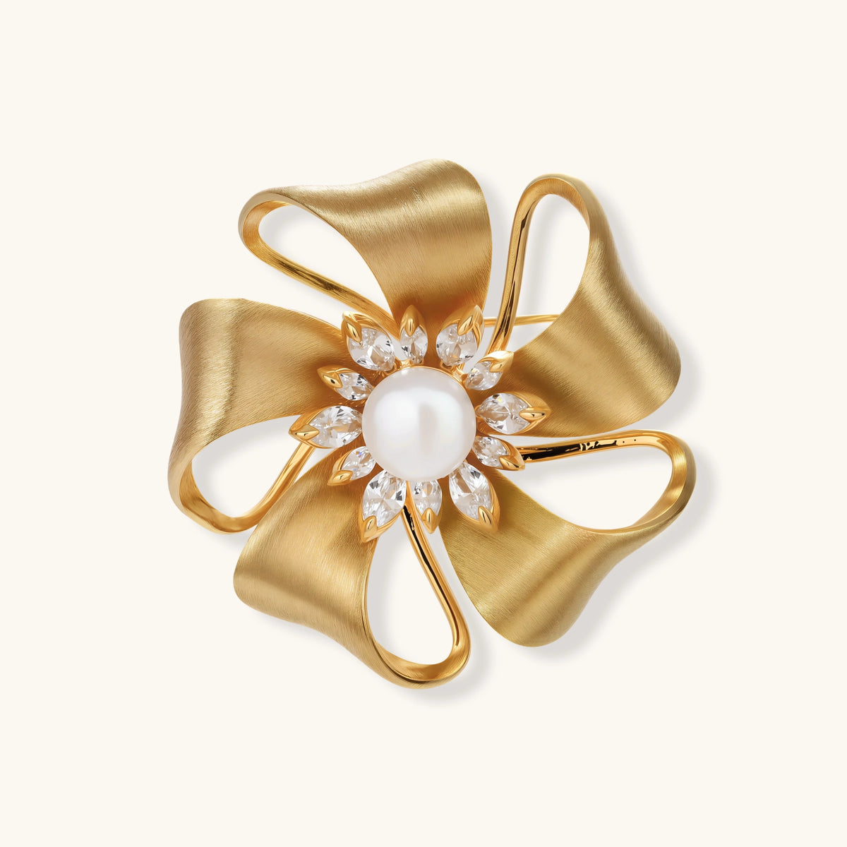 Gold ribbon-petal flower brooch with a round white bead center ringed by marquise zirconia stones on a cream background.